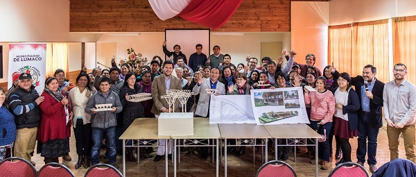 Cerca de 60 feriantes, hombres y mujeres de la comuna de Lumaco, en la primera Reunión Ampliada sobre la Construcción del Mercado Turístico de Lumaco. Tres personas sostienen maquetas de madera. Hay una mesa con maquetas ye imágenes del proyecto