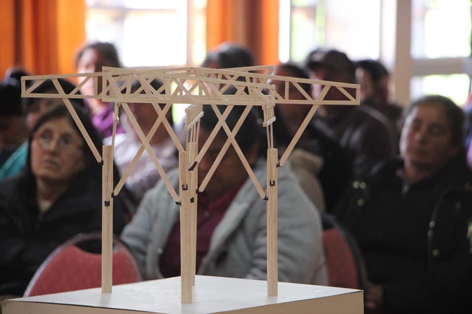 En primer plano hay una maqueta de una estructura de madera. Al fondo personas sentadas en un auditorio 