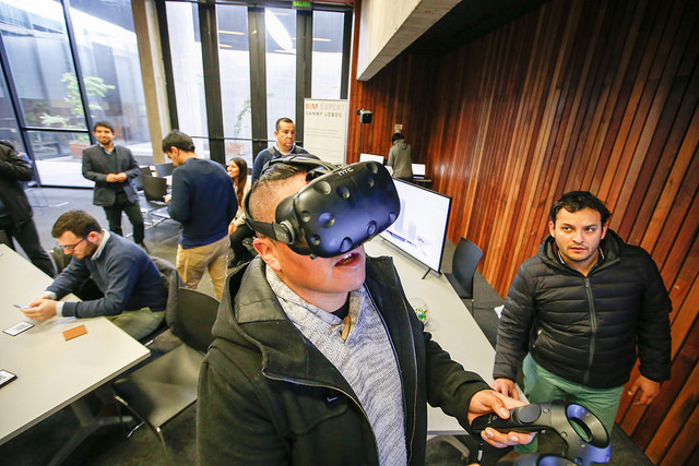 Un hombre con unas gafas de realidad virtual y un control en la mano. De fondo varias personas caminando