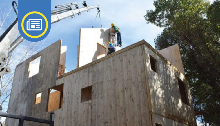 Construyendo una vivienda con paneles de madera. Una grúa sostiene un panel mientras dos trabajadores la posicionan en su lugar